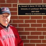 An elderly man wearing a red shirt an black baseball hat standing in front of a brick wall and plaque