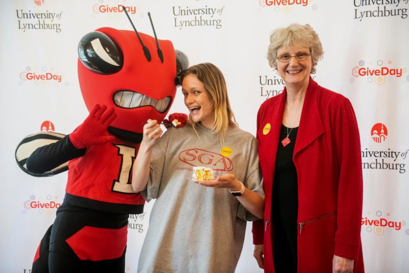 A red Hornet mascot and a smiling female student with long blond hair and an older woman with short gray hair and glasses pose in front of a white backdrop