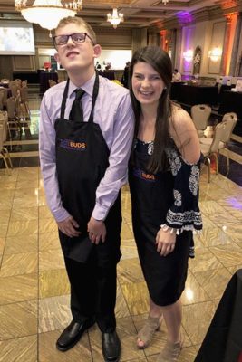 A young white man in glasses, a purple shirt and black tie wearing a black apron poses with a young white girl with long dark hair in a black apron and a sleeveless shirt and sandals, smiling