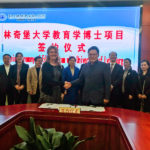 Sally Selden shakes hands with Ziaofeng Wei as others look on in the background. The agreement they signed for their schools sits on a table in front of them.