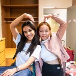 Two female students sitting in a dorm room making a heart with their arms