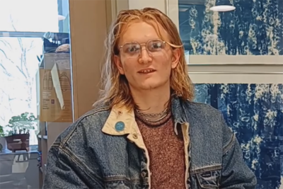 A young white man with long blond hair and glasses wearing a blue denim jacket