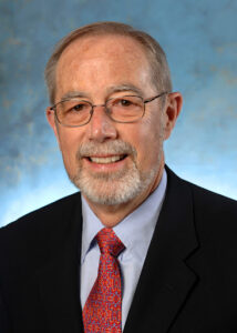 An elderly man with short gray hair and a gray beard and glasses wearing a suit, smiling
