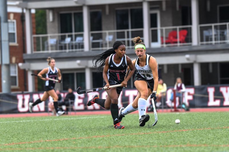 A young Black woman with long braids, wearing a black sleeveless shirt and skirt and carrying a hockey stick runs next to a young white woman with brown hair, wearing a white, sleeveless shirt, black skirt and carrying a hockey stick chasing a ball