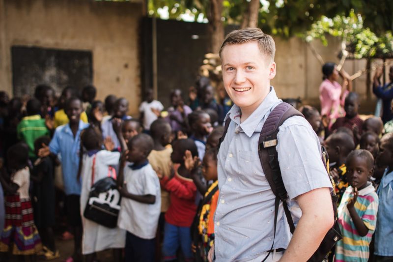 A young white male in a short-sleeved button-up shirt carrying a backpack stands in front of a group of African children, smiling