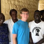 A young white male in glasses and a blue shirt poses with a group of African adults