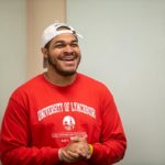 A black male student in a red sweatshirt and a white baseball cap laughing