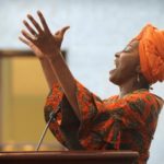Black woman in orange-colored traditional African attire singing and raising her hands
