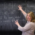 Woman standing in front of and gesturing towards writing on a chalkboard.