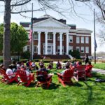 A group of people sit in a circle on red Adirondack chairs on a green lawn under trees in front of a brick building with white columns on a sunny day