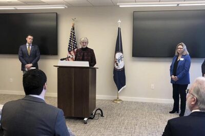 A white woman with short white hair and glasses speaks at a podium. Behind her are two black TV screens and the American flag and the Virginia flag. To her right stands a white man in a black suit, to her left a white woman in a blue suit.