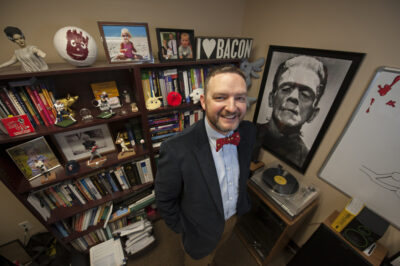 A middle-aged man with short dark blond hair in a suit and bowtie stands indoors in front of a bookcase and a black-and-white painting of Frankenstein's monster