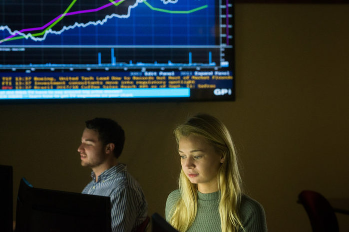 Rebecca Taylor works at a computer in a trading room with a stock market graph on a screen in the background.