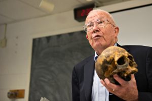 Photo of Dr. William Bass holding a skull. A chalkboard is in the background.