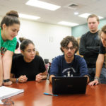 students gather around a table discussing something as one types on a laptop