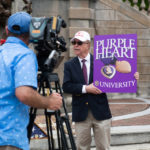 President Garren holding a large purple sign with the words "Purple Heart University" as he talks to reporters