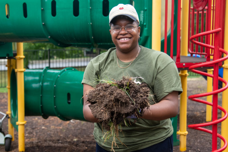 A Black student in a green shirt outside, holding a bundle of weeds and dirt, smiling