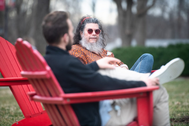 A middle-aged man with sunglasses and a white beard sitting outdoors in a red Adirondack chair talking with a younger, dark-haired man who has his back to the camera