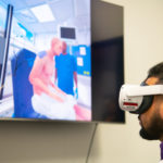darkskinned male wearing a VR headset in front of a TV screen that shows a patient sitting on a blue chair