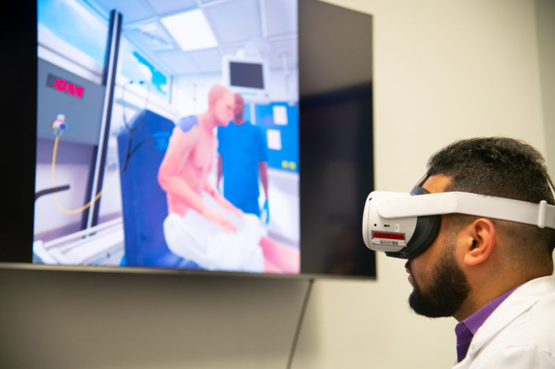 darkskinned male wearing a VR headset in front of a TV screen that shows a patient sitting on a blue chair