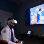 A male student wearing a VR headset in front of a screen that simulates a PA taking care of a patient