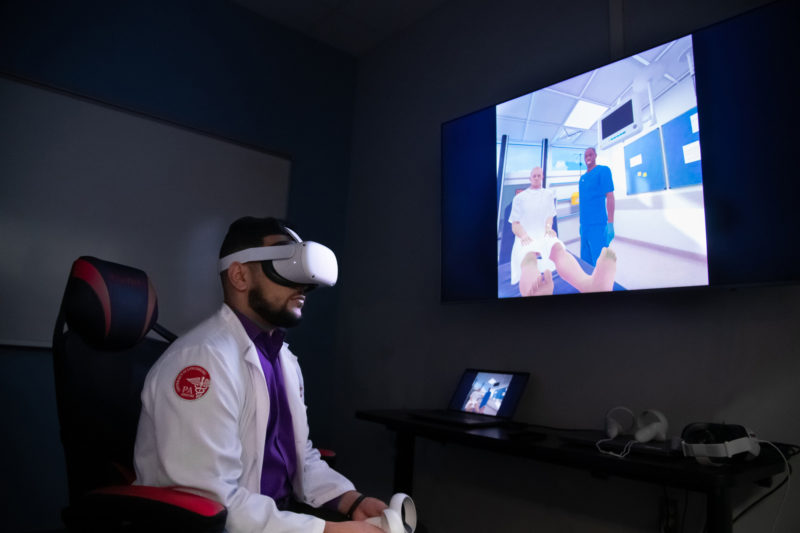A male student wearing a VR headset in front of a screen that simulates a PA taking care of a patient