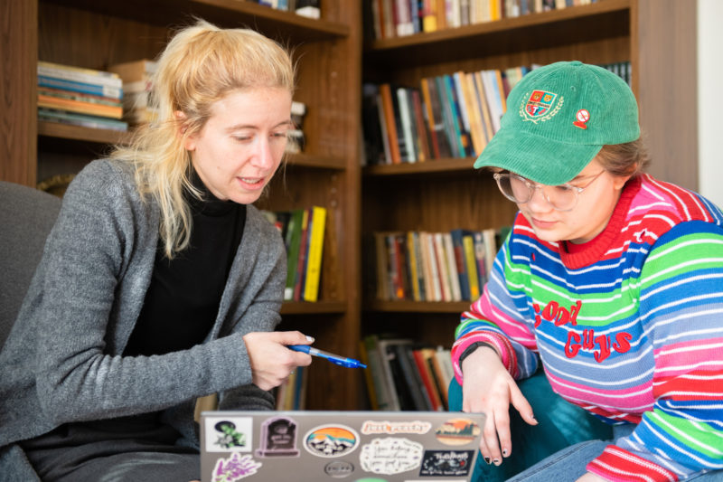 A white woman with long blonde hair dressed in gray and black sits with her laptop covered in stickers and a white female student in a rainbow sweater and baseball hat