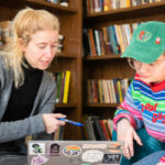 A white woman with long blonde hair dressed in gray and black sits with her laptop covered in stickers and a white female student in a rainbow sweater and baseball hat