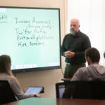 Middle-aged white man standing in front of a smart board teaching a class to a group of students seated with their backs to the camera