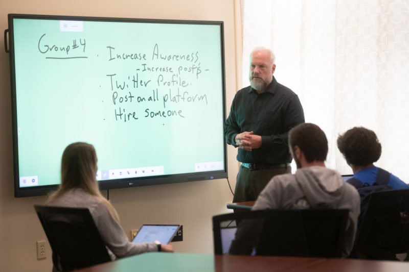 Middle-aged white man standing in front of a smart board teaching a class to a group of students seated with their backs to the camera