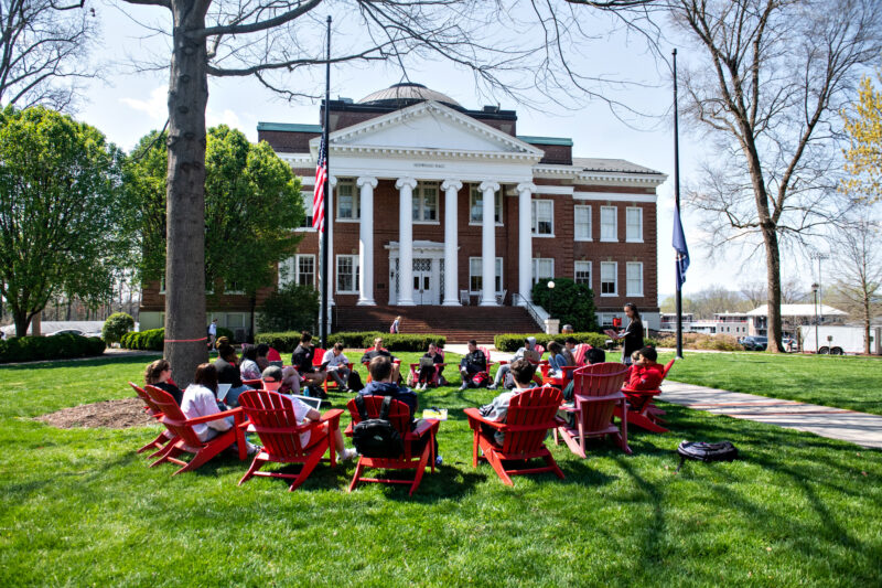 A group of people sit in a circle on red Adirondack chairs on a green lawn under trees in front of a brick building with white columns on a sunny day