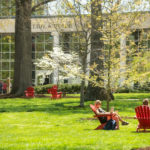 students in red chairs outside on a sunny day with trees and an academic building