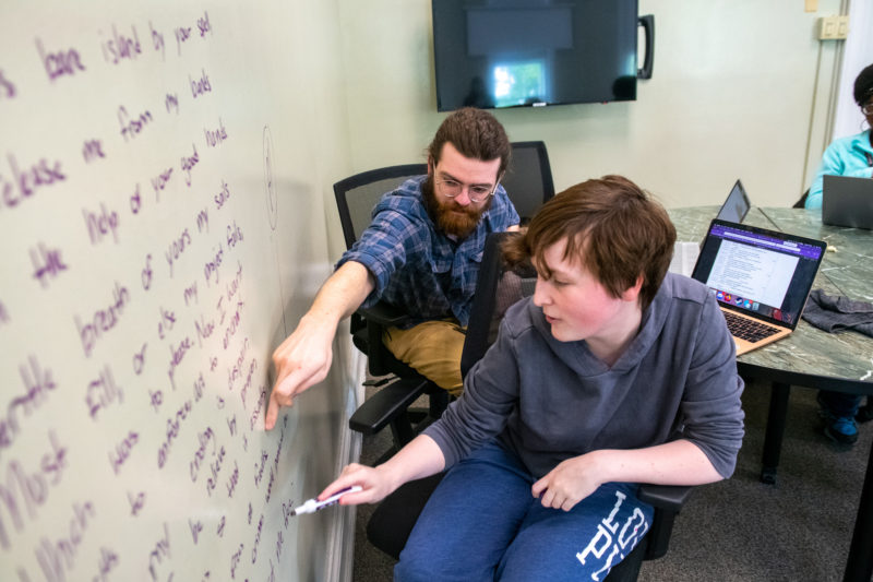 A student in casual clothing and a professor write on a whiteboard