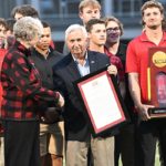 An elderly man in khakis and a black blazer with short white hair holding a frame and a woman with short gray hair in black slacks and a black and red fleece jacket shake hands next to a young man in a red polo holding a trophy surrounded by other young men in athletic clothing