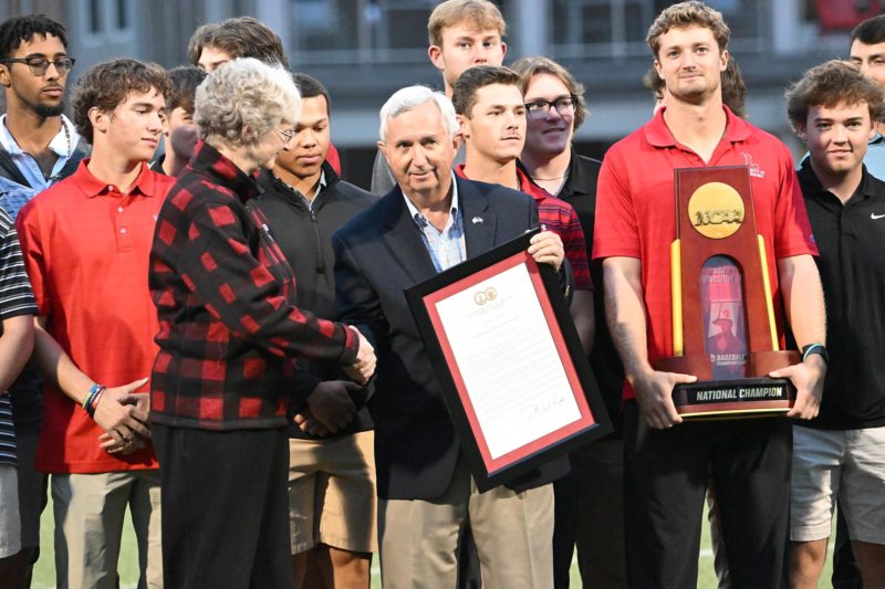 An elderly man in khakis and a black blazer with short white hair holding a frame and a woman with short gray hair in black slacks and a black and red fleece jacket shake hands next to a young man in a red polo holding a trophy surrounded by other young men in athletic clothing