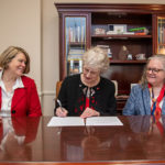 Nancy Hubbard smiles as President Alison signs a piece of paper. Provost Allison Jablonski is seated beside them.