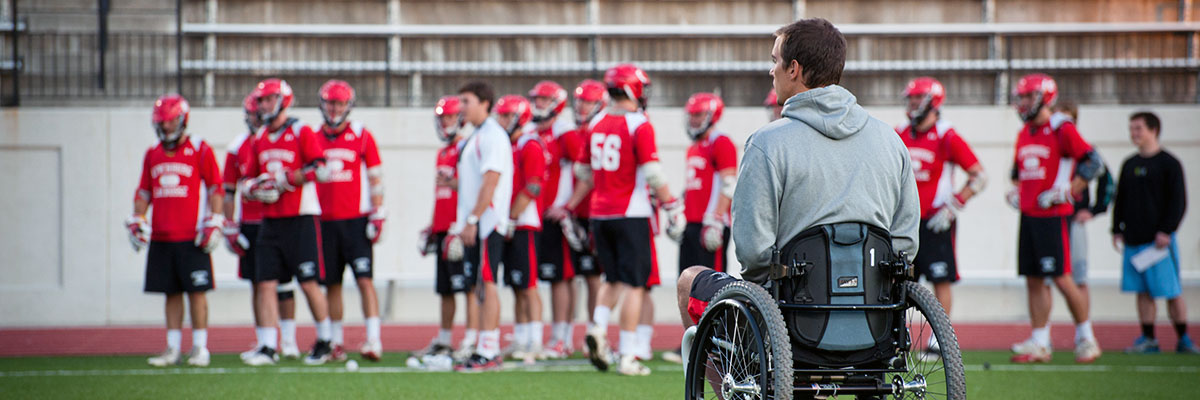 A student in a wheelchair on a lacrosse field with players in the background