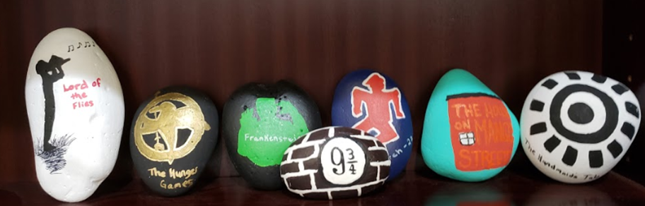 several rocks with book titles or images painted on them are lined up on a bookshelf.