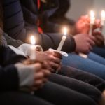 Several people in black and blue jeans and dark coats sit in a pew holding candles
