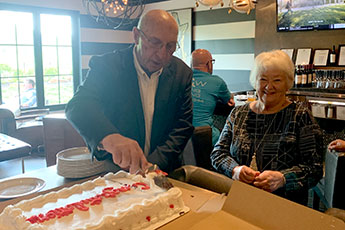 A man cuts a large cake with the word "Congratulations" with his wife by his side.