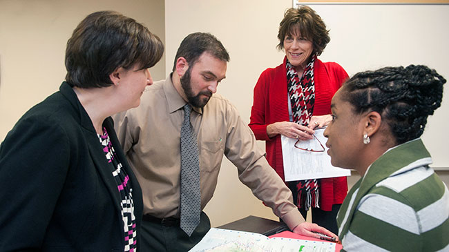Three women and a man gather around a desk consulting paperwork.