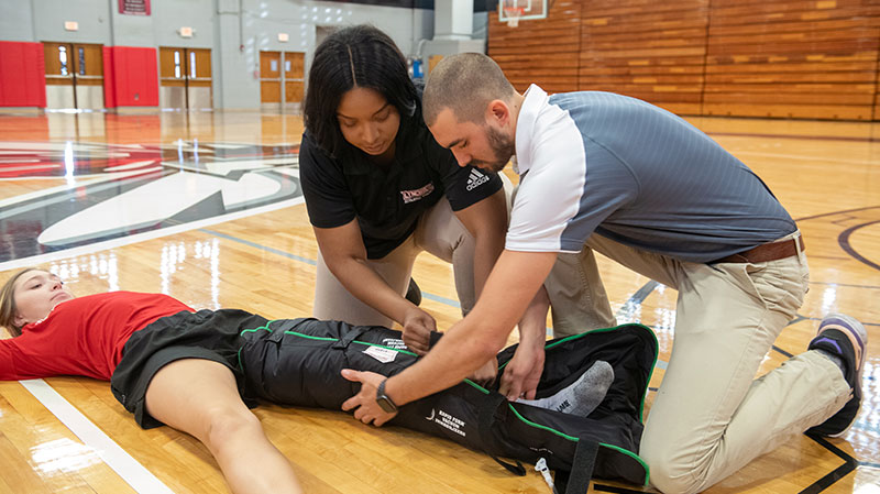 An athlete lying on the floor of a gym with two athletic trainers tending to her leg