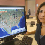 A brown-skinned woman with shoulder-long black hair sits at a computer showing a map