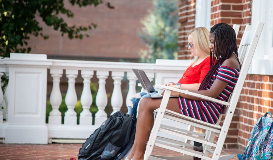 Two students sit in white rocking chairs on a brick porch, working on laptops with backpacks beside them.