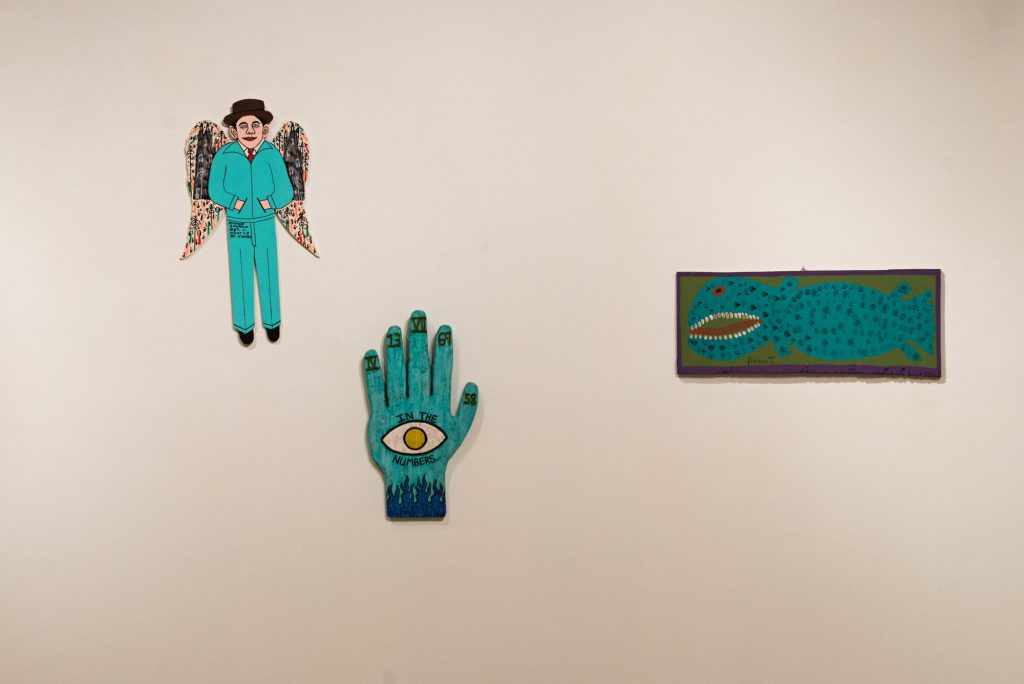 Art work by Howard Finster shows a man with wings and a top hat, and a hand. A nearby painting shows a toothy fish.