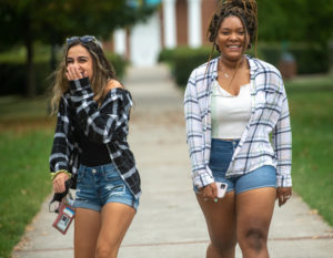 Two students walking on the Dell. One is smiling, and the other is covering her mouth laughing.