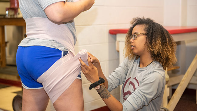 A student wraps an ace bandage around the waist and thigh of another student.