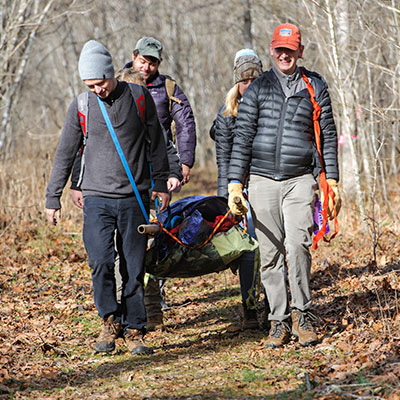 Six students carry supplies on a path through the woods.