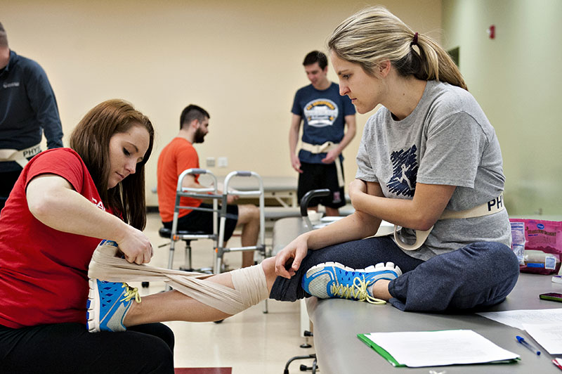 A student sits on a PT table with one leg extended, while another student is bandaging her leg