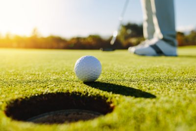 Golf ball sits near the hole on the green as golfer shoes and a golf club are seen in the background.